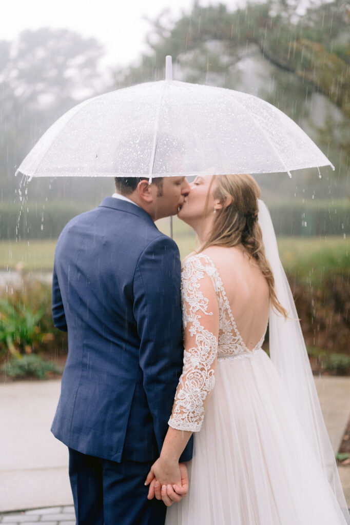 Bride and Groom kiss under an umbrella while the rain is pouring around them.