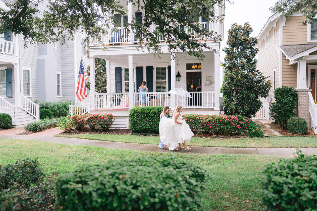 Bride walks across the rainy sidewalk while bridesmaid carries her train and an umbrella.